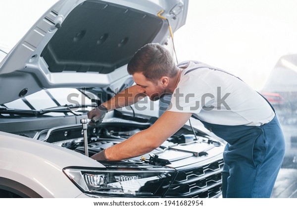 Quality service. Young man in white shirt and\
blue uniform repairs\
automobile.