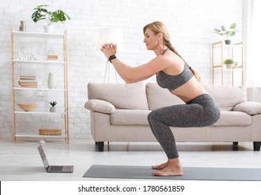 Quarantine and exercise at home. Slim adult woman in sport uniform with fitness tracker crouches and watches at laptop in living room interior, copy space