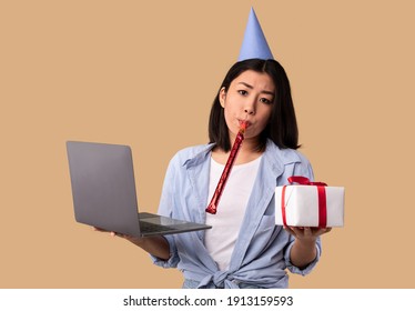 Quarantine, Holidays And Virtual Event Concept. Portrait Of Sad Asian Woman Wearing Hat, Blowing Party Horn Pipe, Celebrating Birthday Alone, Holding Computer And Wrapped Present Box At Studio