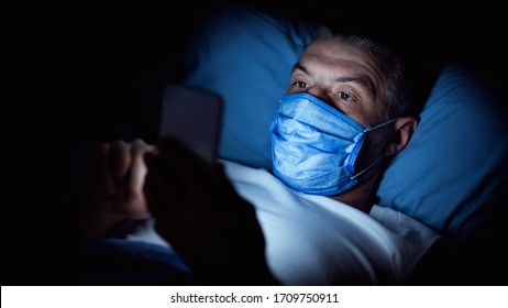 Quarantine Lifestyle. Man Using Mobile Phone Wearing Medical Mask Lying Overnight In Bed At Home. Panorama, Selective Focus