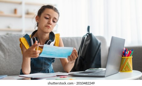 Quarantine And Lockdown Concept. Portrait Of Sad Teenage Schoolgirl Holding Disposable Medical Face Mask, Studying And Learning At Home Using Her Laptop Computer. Upset Bored Girl Sitting At Desk