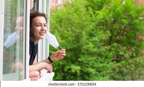 Quarantine is over concept. Young smiling woman taking off medical protective mask looking out of window of her apartment. Happy girl breathing fresh summer air. Freedom, end of coronavirus.Copy space