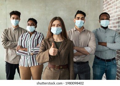 Quarantine, Return To Work Concept. Confident Multicultural Businesspeople Standing In Office With Folded Arms, Wearing Protective Medical Masks, Ceo Showing Thumb Up Gesture, Posing Looking At Camera