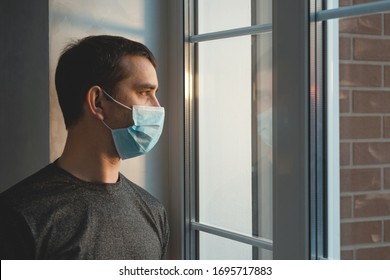 Quarantine self-isolation. Sad young man in a medical mask who looks out the window through the window. Infected man in medical mask on self-isolation looks at the street through the window of a house