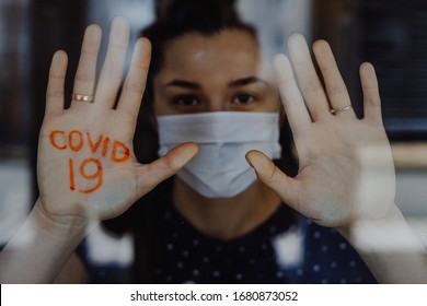 Quarantined Young Woman At Home Looking Out The Window With The Inscription On Her Palm: «covid 19» Meaning Warning Of Coronavirus Infection