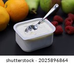 quark or cream cheese in plastic box with spoon on black backround with fruits, healthy nutrition for breakfast