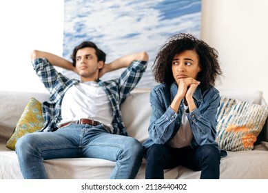 Quarrel, disagreement between a mixed race young couple. A guy and a girl, offended at each other after a quarrel, are sitting on the couch, do not talk to each other - Shutterstock ID 2118440402