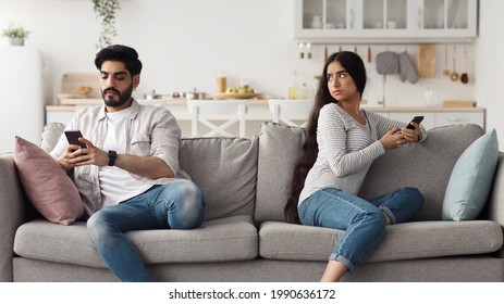 Quarrel, relationship problems, jealousy, suspecting infidelity and gadget addiction. Sad millennial arab wife with gadget looks at husband with phone in living room interior, panorama, copy space