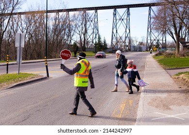 Quebec City, Quebec, Canada, May 11, 2021 - School Crossing Guard Holding Stop Sign Helping Young Smiling Pregnant Mother And Little Girl At Pedestrian Crossing In The Cap-Rouge Area