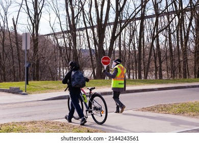 Quebec City, Quebec, Canada, May 11, 2021 - School Crossing Guard Holding Stop Sign Helping Young Boy Walking With His Bicycle At Pedestrian Crossing In The Cap-Rouge Area