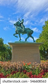 QUEBEC CITY, CANADA - SEPTEMBER 17, 2016: A Bronze Statue Of Joan Of Arc Stands As A War Memorial In A Colorful Garden Under A Blue Sky.