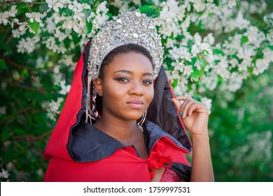 Queen In The Crown And Royal Mantle. Lovely Girl On The Background Of A Blooming Garden. African American In Red