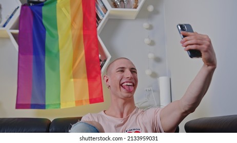 Queer Caucasian Bald Gay Man Sitting In Front Of Rainbow Pride Flag And Taking Selfie Of Himself With His Tongue Out. High Quality Photo