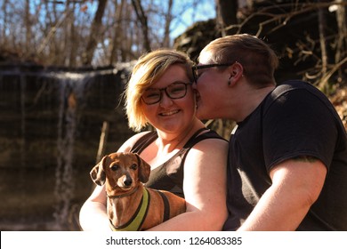Queer couple with lesbian and transsexual male snuggles with their dog near a waterfall