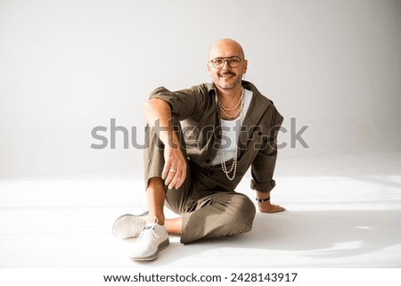 A queer gay man sits on the ground with his legs crossed in a studio fashion portrait. He appears confident and relaxed in his pose, emanating a sense of strength and self-assuredness.