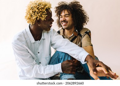 Queer Lovers Smiling At Each Other. Two Young Male Lovers Looking At Each Other Affectionately While Sitting Together Against A Studio Background. Young Gay Couple Sharing A Romantic Moment.