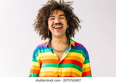 Queer man smiling cheerfully in a studio. Young gay man celebrating gay pride while standing alone against a white background. Young gay man wearing a shirt with rainbow colours.