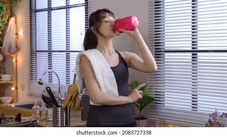 quenching thirst after morning yoga routine. cheerful lady wearing sport outfit with towel on shoulder is enjoying cityscape view from home. healthy lifestyle and real moments