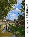 Queralt bridge over the Meder river, 11th century, Vic, Osona, Barcelona, Catalonia, Spain with calm waters and green meadows with a blue sky with clouds and tree branches on its sides.