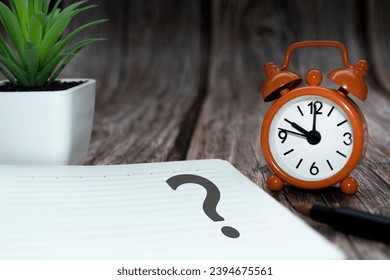 Question mark on notepad alarm clock set at 10 o'clock on wooden background. Q  A concept.