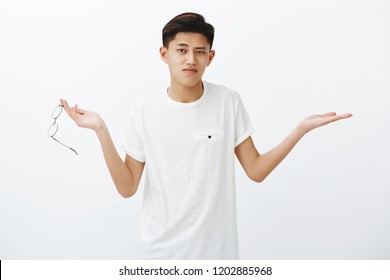 Questioned Asian Guy Cannot Understand What Problem. Portrait Of Confused Shrugging Chinese Male Student Taking Off Glasses Spread Hands Sideways In Clueless Gesture Over White Background