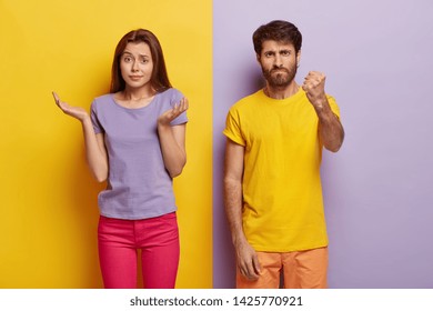 Questioned Doubtful Woman Spreads Palms Sideways, Has Unaware Facial Expression, Angry Man Shows Fist At Camera, Asks Not Bother Him, Pose Indoor Over Colorful Background. Hesitation And Anger