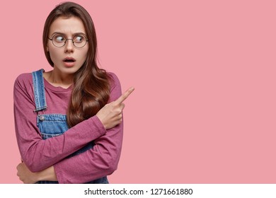 Questioned Woman With Surprised Expression Points Aside With Index Finger, Opens Mouth From Amazement, Watches Amazing Show, Feels Impressed, Isolated Over Pink Background, Can Not Say Word.