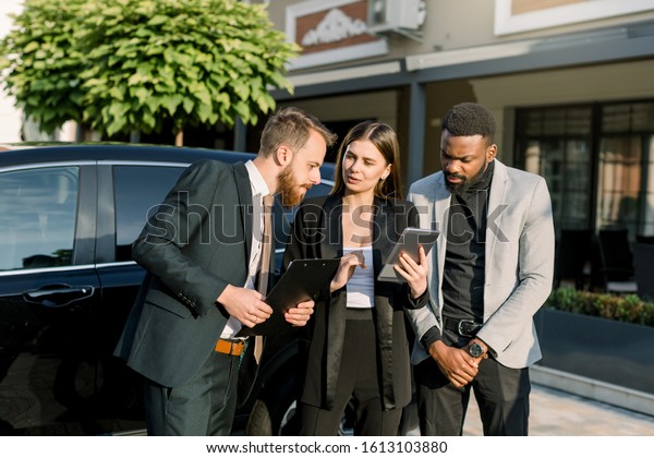 Quick briefing before meeting. Three cheerful\
young multiethnical business people, two men and one woman, talking\
to each other while standing outdoors near the black car. Woman\
holds digital tablet