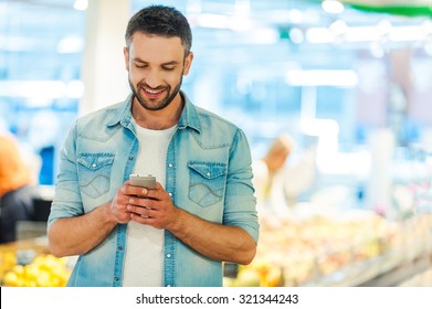 Quick Text During Shopping. Handsome Young Man Holding Mobile Phone And Smiling While Standing In A Food Store 