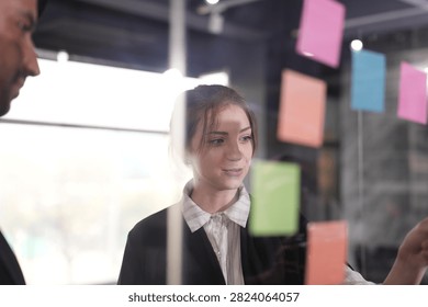 Quiet consideration during the sticky note session, the businesswoman with a bun reading suggestions while her bearded colleague hovers at the very edge of the frame.