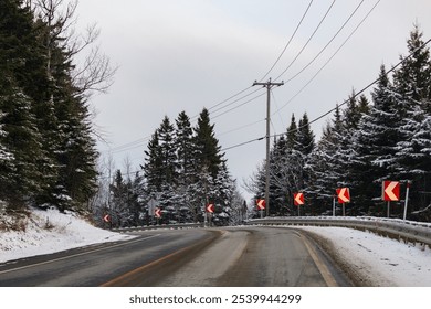 A quiet, empty road winds through a snow-covered forest, adorned with directional traffic signs. Overhead power lines hint at remote tranquility in the peaceful winter landscape. - Powered by Shutterstock