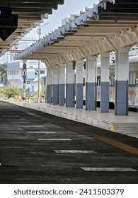 a quiet and empty train station platform, with a series of benches and a roof providing shade, while the tracks lay bare without any trains. The platform is covered by a roof