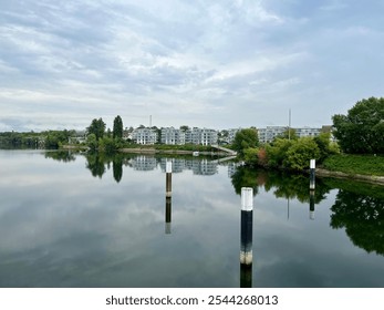 A quiet riverside scene with modern apartment buildings reflected in still water, surrounded by greenery and an overcast sky. - Powered by Shutterstock