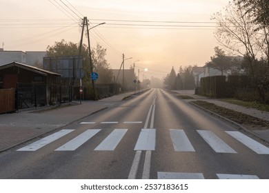 Quiet suburban road with pedestrian crossing under misty morning sunrise, lined with trees and residential buildings. Peaceful urban environment, early morning commute, and traffic infrastructure.  - Powered by Shutterstock