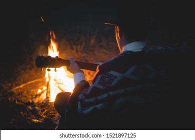 Quiet tranquil time in the night by the campfire, soft focus. Man in traditional native american poncho and hat plays the guitar by the fire in the wood
