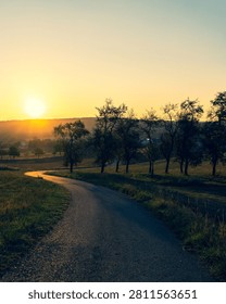 Quiet walking path winding through open fields and trees as the sun sets over a peaceful rural landscape.