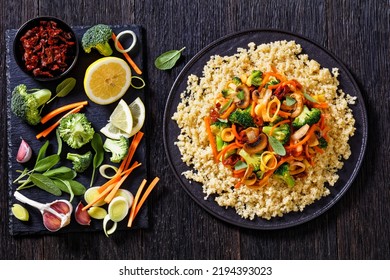 quinoa topped with stir fried broccoli, julienne carrots, sun dried tomatoes, leek and mushrooms on black plate on dark wooden table with ingredients, horizontal view from above, flat lay