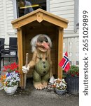 A quirky Norwegian happy troll statue stands proudly bellow a small wooden house surrounded by flowers and a small Norwegian flag.Traditional norwegian troll welcomes visitors.Scandinavian symbol