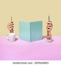 Quirky office concept with upright open notebook flanked by a mans hands holding pen and pencil with head obscured behind the pages , a coffee cup and flower with copy space on a two tone background