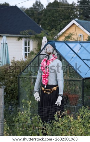 A quirky scarecrow dressed in a sweater and skirt stands guard beside a greenhouse in a lush garden.