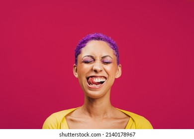 Quirky and vibrant. Excited young woman sticking her tongue out with her eyes closed while standing against a pink background. Fashionable young woman wearing makeup with purple hair in a studio. - Shutterstock ID 2140178967