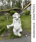 Quirky white stone troll statue holding a wooden staff, set in a lush green outdoor setting with rocks and trees in the background.