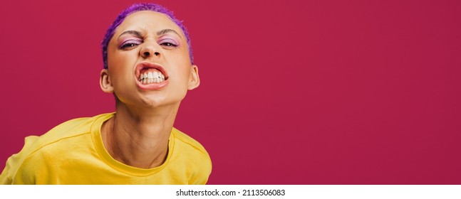 Quirky young woman making a face in a studio. Fashionable young woman looking at the camera confidently while standing against a pink background. Stylish young woman wearing makeup with purple hair.