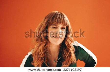 Quirky young woman winking at the camera while standing in a studio. Young woman with ginger hair standing against a vibrant orange background in casual clothing.