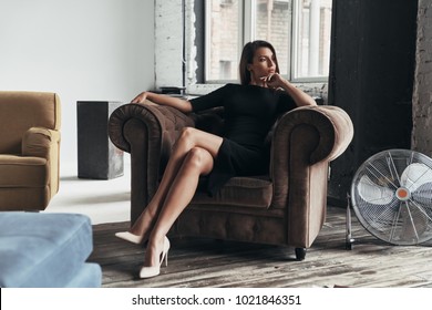 Quite contemplation. Attractive young woman in elegant black dress keeping hand on chin and looking away while sitting in the armchair