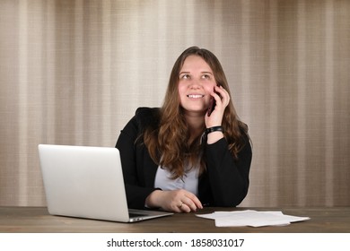Quite dreamy female office worker busy with her thoughts while sitting at the desk in front of laptop and some papers. She wears black strict suit. Her hair is brown. Light & dark brown background. - Shutterstock ID 1858031017