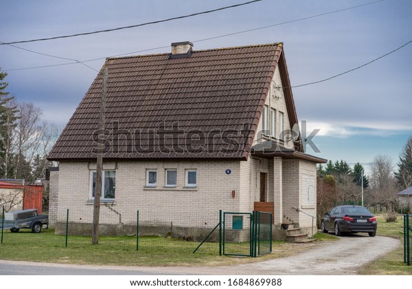 Raasiku / Estonia - March 27 2020: Residential\
neighborhood in rural parish. Small simple two storey detached\
brick house with pitched roof, green wooden fence. Black new BMW\
parked next to the\
house.