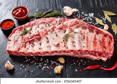 rack of raw pork spare ribs seasoned with spices on black slate tray with paprika, garlic cloves, bay leaves at background, view from above, close-up