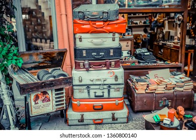 Racked Suitcases In Front Of Antique Shop. Colorful Old Suitcases. Old Plaques And Books