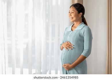 radiant Asian pregnant woman stands near a window draped with sheer curtains, gently cradling her belly while looking out with a content smile. - Powered by Shutterstock